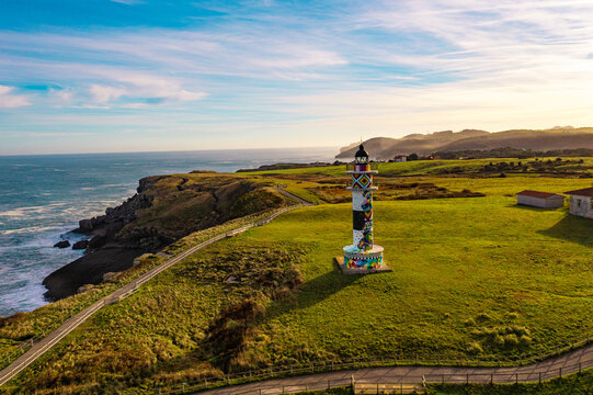 Colorful Ajo Lighthouse captured by drone, standing on a green cliff with a vast ocean backdrop. Coastal landscape of Faro de Ajo in Cantabria, Spain