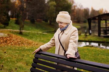 Sad elderly woman standing alone in autumn park, looking down with thoughtful expression. Depressed senior female outdoors surrounded by golden trees and fallen leaves, symbol of loneliness and aging