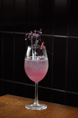Pink gin and tonic cocktail, presented in a clear glass with delicate flowers and a red clothespin, rests on a wooden surface with dark tiles behind. The blend of gin and tonic creates a fizzy