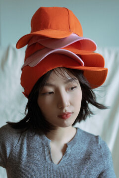 Young Asian woman wearing multiple colorful hats stacked on her head in a playful indoor setting, showcasing her creative style