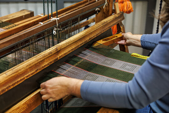 Close-up of a person weaving fabric on a wooden loom with green, grey, and red threads. A yellow measuring tape lies nearby. Traditional textile craft scene