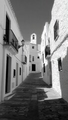 Monochrome street scene with white buildings, dark doorways, balconies, and a steeple in the distance, showcasing a narrow, paved road.