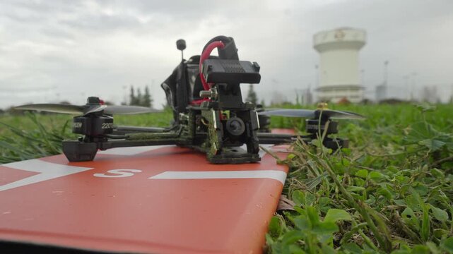 The drone sits among blades of grass while the arm switch is engaged, its motors gaining subtle momentum before launch.