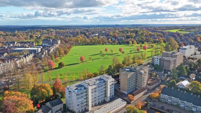 Drone view of Harrogate, North Yorkshire, England, showing autumn trees surrounding the large open parkland known as The Stray, with residential and historic town buildings under a blue clody sky.