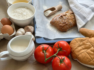 Fresh-baked bread on baking sheet with oven glove, flour, milk, heads of garlic, eggs, tomatoes 