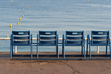 Four iconic blue chairs lined up on the famous promenade in Nice, France, overlooking the calm Mediterranean Sea with yellow buoys. © Dmitri