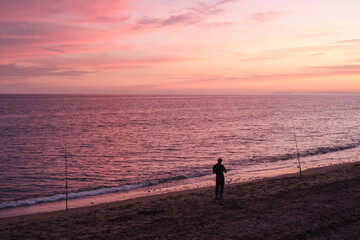 Fototapeta premium Fisherman silhouette at sunset on a quiet beach with pink sky and gentle waves, peaceful seaside scene for travel and lifestyle concepts.