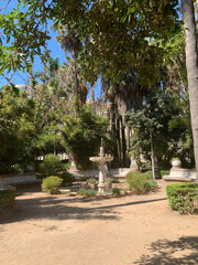 A small, elegant carved fountain in the courtyard of a public space with a surrounding garden. Spain, 2022.