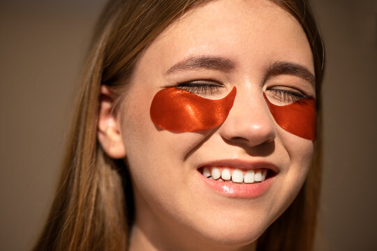 Young woman with long hair smiles while wearing under-eye patches, showcasing a skincare routine, illuminated by soft natural light, emphasizing beauty and self-care