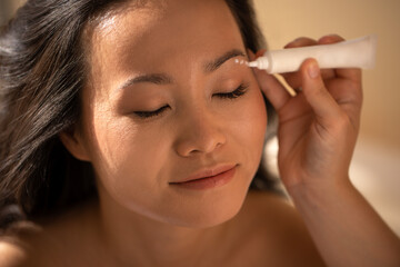 Obraz premium Asian woman receiving skincare treatment, applying cream to her face, with soft lighting enhancing her natural beauty, showcasing self-care and relaxation in a serene environment
