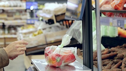 Shopper uses a digital scale to weigh a plastic bag of fresh bell peppers in the produce section of a supermarket before making a purchase