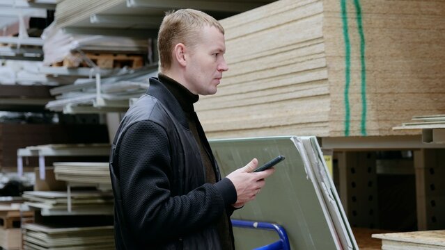 Construction worker checking inventory on smartphone in lumber warehouse surrounded by building materials. A man in a hardware store, choosing lumber, shelves with lumber.
