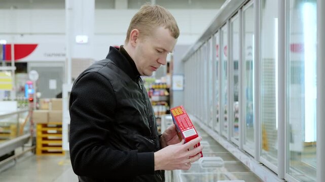 Customer choosing products in supermarket trying to make a decision.. Customer wearing a black jacket is reading a frozen food label at a supermarket. He is standing in the frozen food aisle
