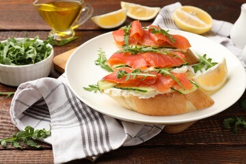 Tasty bruschettas with salmon, cream cheese, cucumber, arugula and lemon on wooden table, closeup