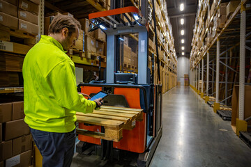 Warehouse employee using a tablet to operate a pallet stacker in a large storage facility. The worker in a high-visibility jacket checks inventory among tall shelves filled with cardboard boxes.