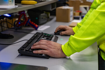 Warehouse employee using a computer keyboard and mouse at a packing station. The worker in a high-visibility jacket manages logistics and inventory tasks in a modern warehouse environment.