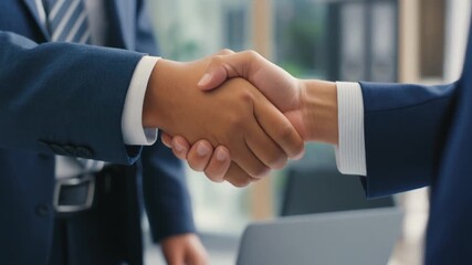 Two businessmen in suits shaking hands in a modern office setting, symbolizing agreement and partnership. - Powered by Adobe