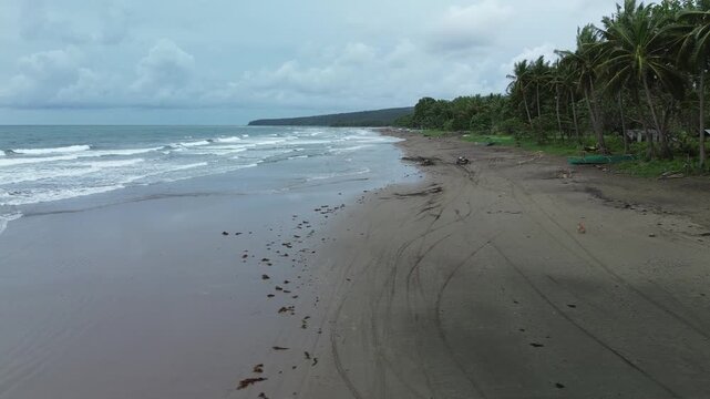 Slow side drift at Little Siargao, Palawan: small onshore waves meet a dark wet beach while coconut palms, a hut, and beached outrigger boats line the coast. Overcast, diffused light.
