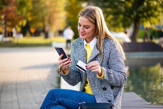 Portrait of modern businesswoman in coat using smartphone and credit card while sitting on a bench in the city park during sunny autumn day.