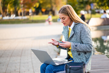 Portrait of urban businesswoman in coat using earphones and laptop while sitting on a bench in the city park. She is talking and gesturing during a video call.