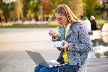 Urban businesswoman with eyeglasses in coat using laptop and enjoying  a healthy lunch break while sitting on a park bench in the city during sunny autumn day.
