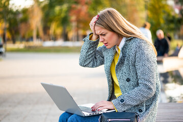 Portrait of overworked and stressed businesswoman with headache using laptop while sitting on a park bench in the city. 