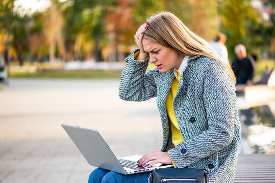 Portrait of overworked and stressed businesswoman with headache using laptop while sitting on a park bench in the city. 