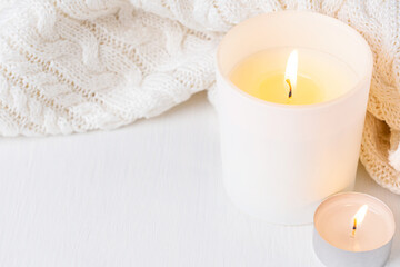 Cozy festive glowing candle in glass jar and knitted blanket or warn sweater on white wooden table with copy space prepared for comfortable evening relaxation at autumn or winter season as decoration
