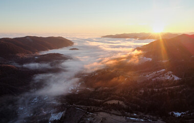 Setting sun casts golden hue over vast sea of clouds, blanketing valleys between forested hills and distant mountain ranges. Aerial view of tranquil scene captures serene beauty of nature.