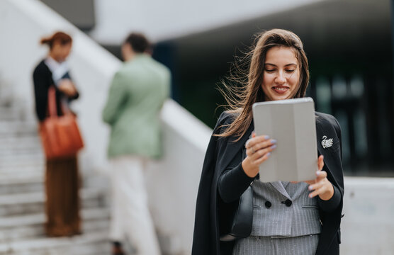 A stylish woman in a blazer and vest uses a tablet outdoors, with blurred colleagues in the background, conveying modern work, connectivity, and friendly collaboration in an urban setting.