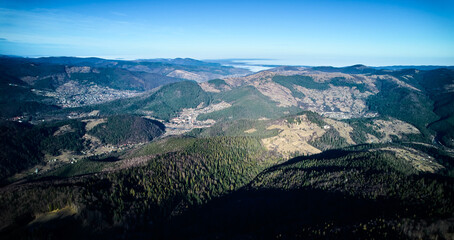 Aerial panoramic view of mountain landscape with village nestled in valley. Rolling hills, forests, sunlit clearings under clear blue sky. Village sprawls out, blending with surrounding landscape.