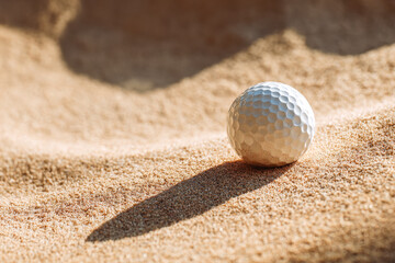 A white golf ball resting on textured sand with a long shadow cast by the sunlight during a clear day on a golf course bunker area
