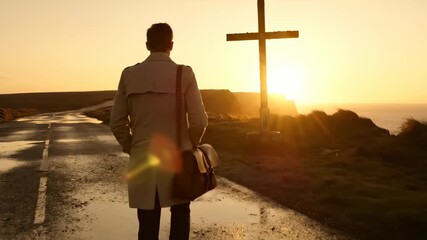 Man on walk toward cross at sunset. Lone figure moves along wet road. Silhouette approaches cross on horizon. Pilgrimage faith in quiet journey. Man carries case toward cross. Golden sunset glow. - Powered by Adobe