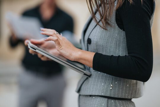 A woman in a gray herringbone vest and black sleeves holds a tablet while a blurred colleague appears in the background, conveying focus and collaboration in a modern workplace.
