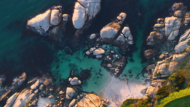Aerial view of the coastline where turquoise waters meet the rugged, sun-kissed boulders of the beach, creating a stunning contrast between land and sea, Cape Town, Western Cape, South Africa.