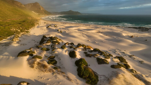 Aerial view of sandy dunes kissed by the sun near the ocean, juxtaposed against rugged mountains, creating a stunning coastal panorama, Cape Town, Western Cape, South Africa.