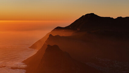 Aerial view of cliffs shrouded in a golden haze meet the ocean's edge under a vibrant sunset sky, Cape Town, Western Cape, South Africa.