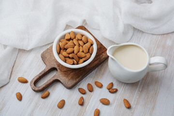 Almond milk in white pitcher and almonds in bowl on wood board, light table background