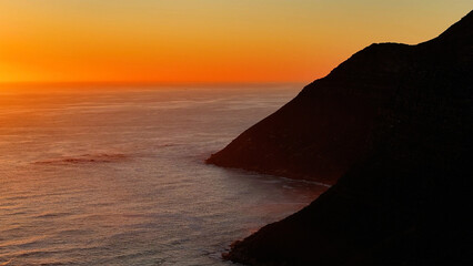 Aerial view of a dramatic coastline where the dark silhouette of the cliffs meets the fiery glow of the setting sun over the vast ocean., Cape Town, Western Cape, South Africa.
