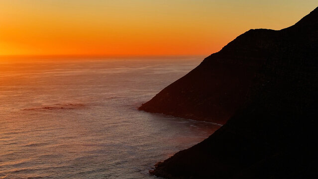 Aerial view of a dramatic coastline where the dark silhouette of the cliffs meets the fiery glow of the setting sun over the vast ocean., Cape Town, Western Cape, South Africa.
