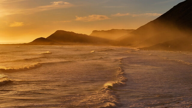 Aerial view of the coastline bathed in the warm glow of the setting sun, where the foamy waves meet the dark, rugged mountains, Cape Town, Western Cape, South Africa.