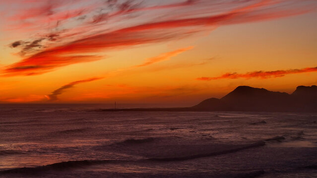 Aerial view of a fiery sunset paints the sky over the tranquil ocean waves meeting the distant silhouette of mountains, Cape Town, Western Cape, South Africa.