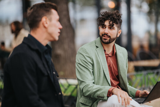 Two men sit outside a cafe, engaged in a relaxed conversation. One wears a green blazer and uses a laptop, while the other listens. A casual, urban setting conveys collaboration and modern work life. - Powered by Adobe
