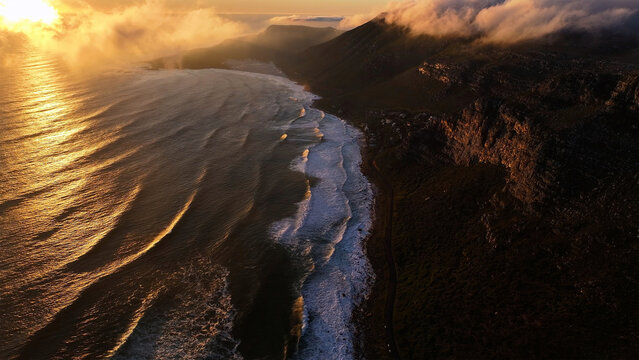 Aerial view of golden light kissing the frothy waves as they meet the dark, rugged coastline near Cape Town, Western Cape, South Africa.