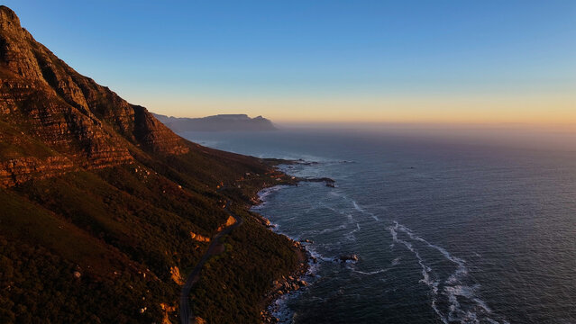 Aerial view of the rugged coastline where the mountains meet the sea, bathed in the warm glow of the setting sun, Cape Town, Western Cape, South Africa.