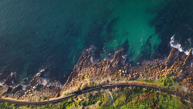 Aerial view of the coastline with turquoise waters meeting rugged rocks and a railway line snaking along the edge, Cape Town, Western Cape, South Africa.