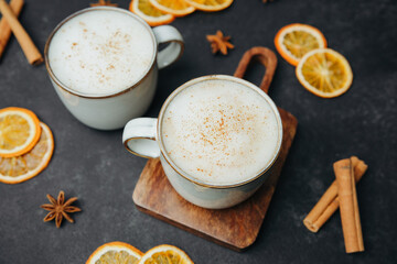 Two warm mugs of frothy drink with cinnamon sprinkle, garnished with star anise, dried oranges, and cinnamon sticks on dark surface