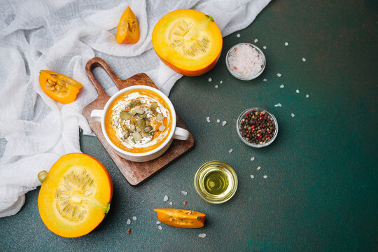 Autumn pumpkin soup with seeds and cream, served in a bowl with fresh pumpkins and spices on a dark table.