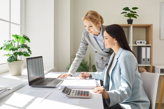 Two smiling businesswomen working together in office, checking spreadsheet on laptop and discussing charts. Female colleagues analyzing financial reports and data, using calculator and paperwork.