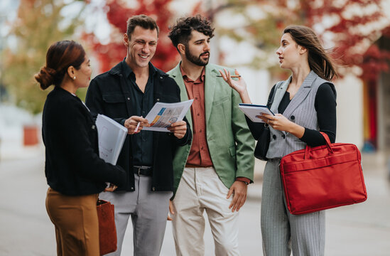A diverse group chats and reviews papers outside on a fall day, sharing ideas and coordinating plans. They wear smart casual outfits and carry bags and folders. - Powered by Adobe